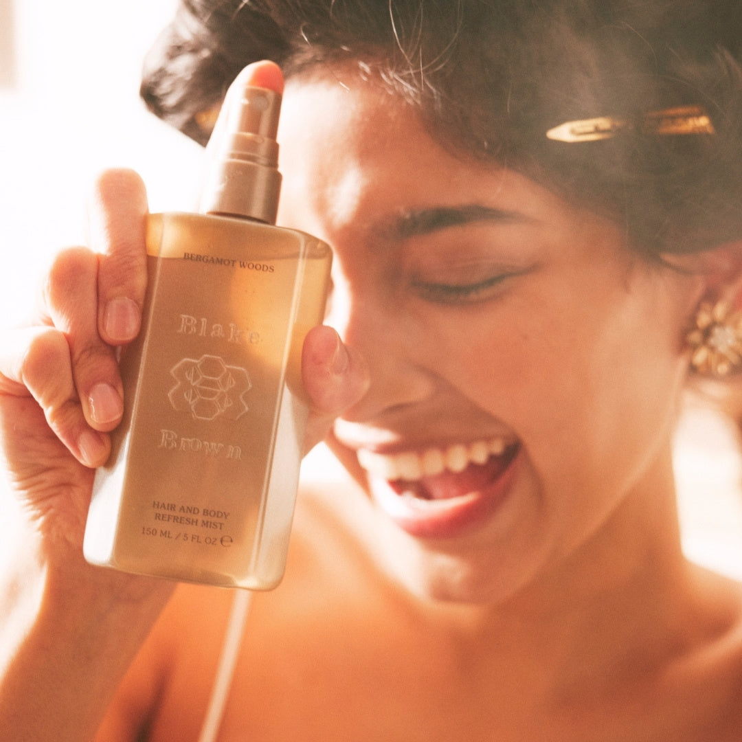 Smiling woman holding a bottle of Blake Brown Hair and Body Refresh Mist in Bergamot Woods scent, mid-spray, with soft golden lighting.