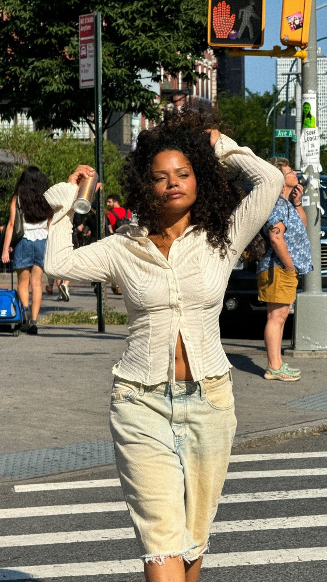 Woman with curly hair walking through a city crosswalk, holding a can of Blake Brown Dry Shampoo.