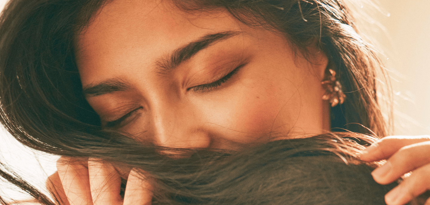 Close-up of a woman with her eyes closed, holding a section of her dark hair near her face, with soft lighting and a serene expression.