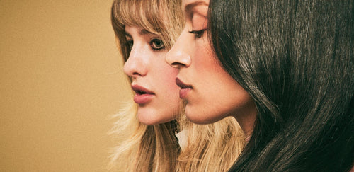 Two women in close-up profile, one with long blonde hair and the other with long straight black hair, both facing left against a warm neutral background.
