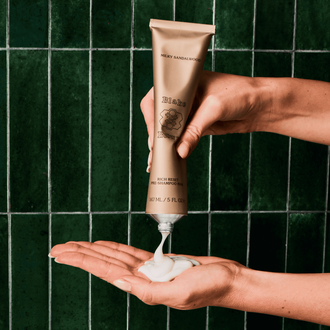 Hands squeezing Blake Brown Rich Reset Pre-Shampoo Mask in Milky Sandalwood into palm against green tile background.