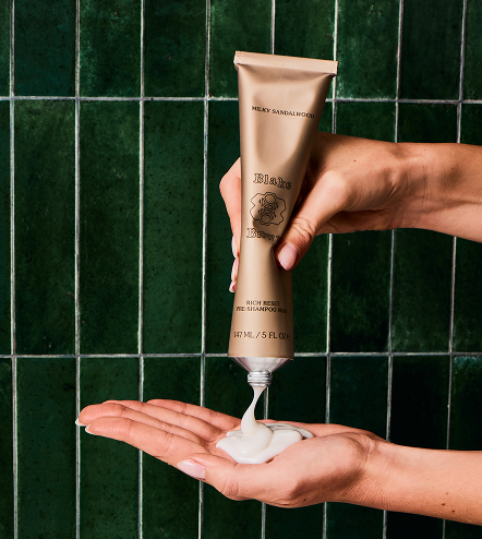 Hands squeezing Blake Brown Rich Reset Pre-Shampoo Mask in Milky Sandalwood into palm against green tile background.