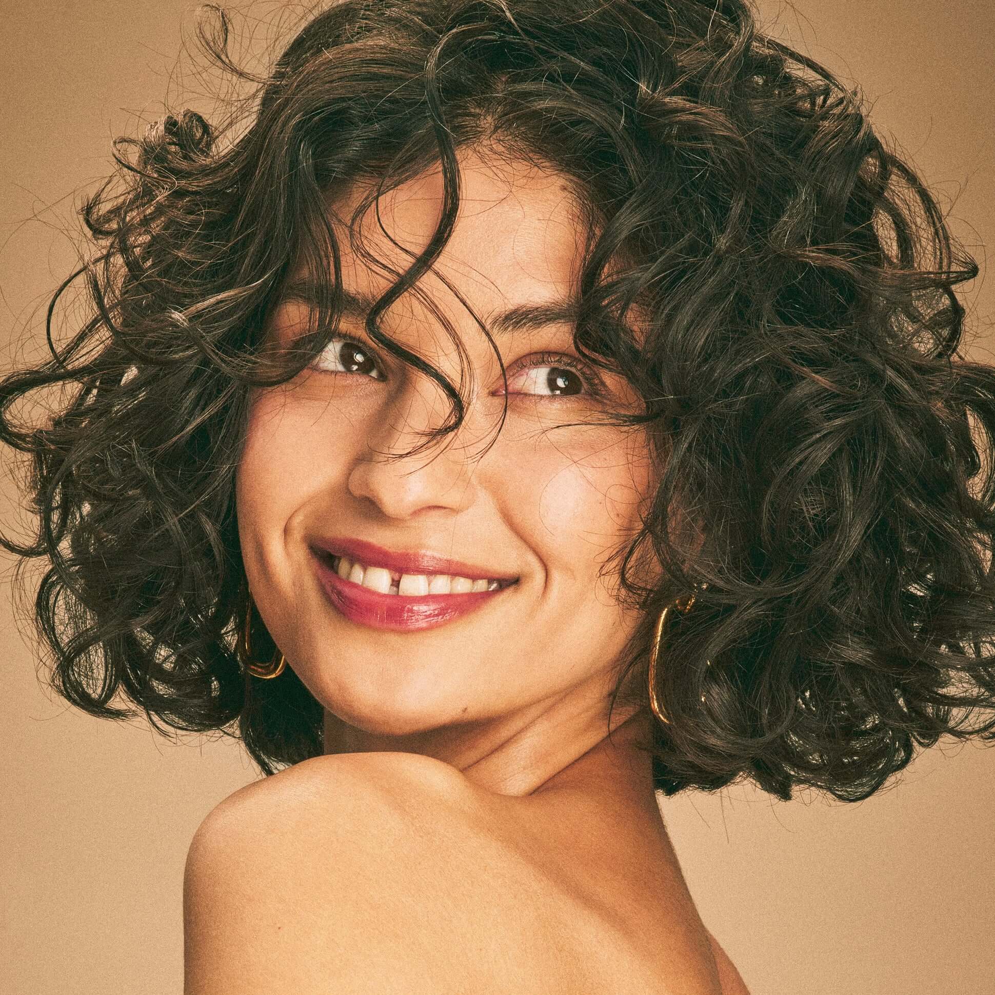 Smiling woman with curly brown hair, highlighting the bounce and shine provided by Blake Brown by Blake Lively's hair-stengthening products.