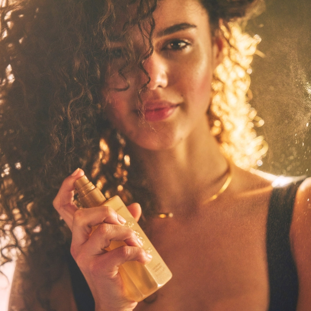 Smiling woman with curly hair spritzing Blake Brown Hair and Body Refresh Mist, surrounded by golden light—capturing a radiant, uplifting self-care moment.