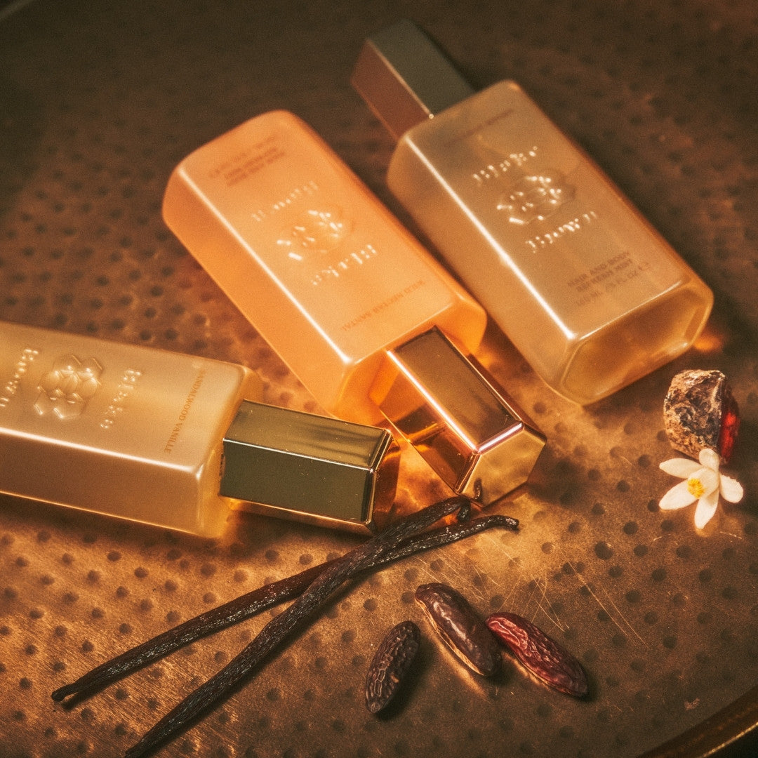 Three Blake Brown Hair and Body Refresh Mists arranged on a warm, textured surface with vanilla beans, tonka beans, and a white flower—evoking rich, warm fragrance notes and a luxurious mood.